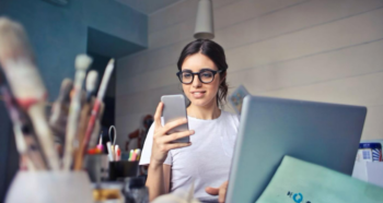 A woman looking at her phone while working on a laptop in a creative workspace