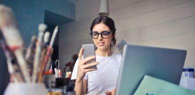 A woman looking at her phone while working on a laptop in a creative workspace